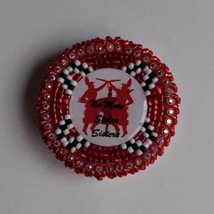 May include: A round, beaded pin with a red and white design. The pin features a central image with two red figures and the text "No More Stolen Sisters". The outer edge is red with clear rhinestones.