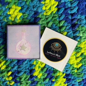 May include: Two stickers, one with a pink potion bottle with a star inside and a purple, blue, and white background, and the other with a black circle with a gold outline and a clock face inside, with the text "Lawrence's Khaos Creative Arts and Mystics" in white.