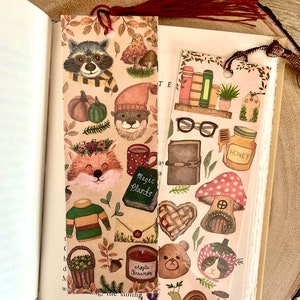 May include: Two decorative bookmarks with autumn-themed illustrations. One features a raccoon, fox, and other woodland creatures. The other has books, glasses, and a honey jar. Both have a tassel and are placed in an open book.