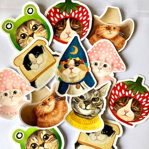 May include: A collection of whimsical cat stickers. Each sticker features a cat wearing a costume, including a frog hat, strawberry hat, cowboy hat, and a wizard hat. The stickers are colorful and detailed.