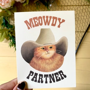 Puede incluir: Tarjeta crema con una ilustraci&oacute;n de dibujos animados de un gato naranja con un sombrero de vaquero. El texto "MEOWDY PARTNER" est&aacute; impreso encima y debajo de la imagen del gato en fuentes marrones y negras.
