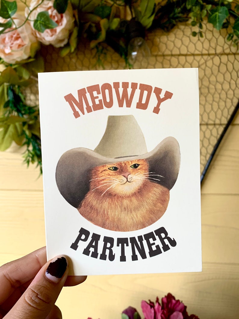 Puede incluir: Tarjeta crema con una ilustraci&oacute;n de dibujos animados de un gato naranja con un sombrero de vaquero. El texto "MEOWDY PARTNER" est&aacute; impreso encima y debajo de la imagen del gato en fuentes marrones y negras.