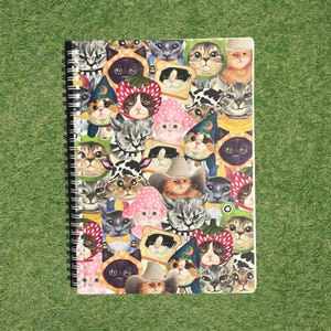 May include: Spiral-bound notebook featuring a collage of cat faces wearing various costumes, including hats, strawberry bonnets, and toast. The cats are in a variety of colors, including gray, orange, and black. The notebook is on a green background.