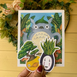 May include: A handmade card featuring a gray Totoro character surrounded by lush green foliage. Additional stickers include a bonsai tree in a yellow pot, a snake plant, and a No-Face character. The card is held against a yellow background.