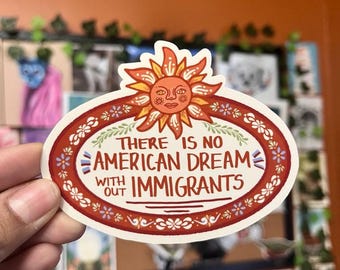 May include: Oval sticker with a sun design at the top and the text "THERE IS NO AMERICAN DREAM WITHOUT IMMIGRANTS" in red and white. The sticker has a floral border and is held by a hand.