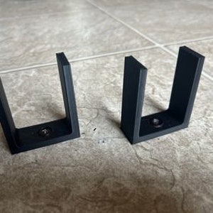 May include: Two black plastic wall mounts with a U-shaped design. Each mount has a screw in the bottom.