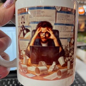 May include: White ceramic coffee mug featuring a colourful illustration of a person seated at a desk with a laptop, surrounded by papers, coffee cups, and other office supplies. The person is wearing glasses and has a stressed expression.