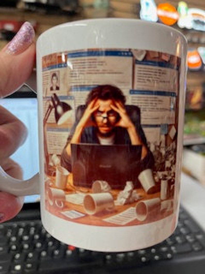 May include: White ceramic coffee mug featuring a colourful illustration of a person seated at a desk with a laptop, surrounded by papers, coffee cups, and other office supplies. The person is wearing glasses and has a stressed expression.