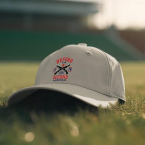 May include: A light gray baseball cap with the text "DEFEND THE SECOND AMENDMENT" in red and blue. The cap features crossed rifles and the numbers "1776". The hat is resting on green grass.