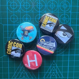Puede incluir: Cinco botones redondos con diferentes diseños. Los botones son negros, azules, rojos y turquesa. Los botones tienen texto que dice "Comic Con", "San Diego Comic Con", "San Diego, CA" y "H".