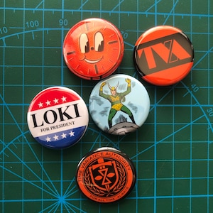 Può includere: Quattro bottoni rotondi con diversi design. Il primo bottone è rosso con un quadrante dell'orologio e un viso sorridente. Il secondo bottone è rosso con testo nero che dice "TA". Il terzo bottone è rosso, bianco e blu con il testo "LOKI FOR PRESIDENT". Il quarto bottone è nero con un cerchio dorato che ha una spada e il testo "TIME VARIANCE AUTHORITY FOR ALL TIME ALWAYS".