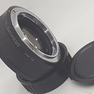 May include: A black teleconverter lens with a silver ring and the text "TELEMORE 0.65 FOR T3" printed on the side. The lens is attached to a black lens cap.