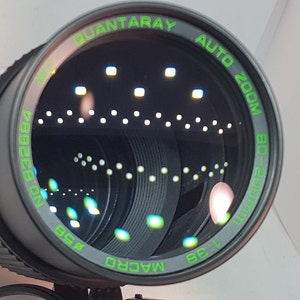 May include: A black camera lens with green text that reads "Quantaray Auto Zoom 200mm 1:3.8 Macro".