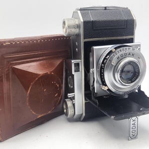 May include: Vintage Kodak camera with a silver lens and black body, accompanied by a brown leather case. The camera features a Compur lens and a Kodak label. The case is rectangular with a geometric design.
