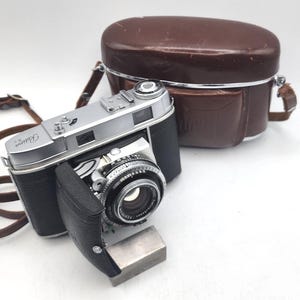 Può includere: Una fotocamera vintage argento e nera con custodia e cinturino in pelle. La fotocamera ha un obiettivo e una copertura protettiva. La custodia in pelle marrone è di forma ovale. Il cinturino della fotocamera è marrone.