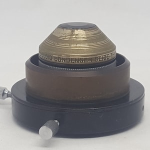 Può includere: Un condensatore per diapositive Polaroid vintage. Il dispositivo ha una base nera con due manopole di regolazione argentate. Una sezione cilindrica marrone supporta una parte superiore dorata a forma di cono con la scritta "POLAROID CONDENSER SLIDE".