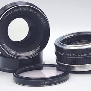 May include: A collection of camera lenses and accessories. The image features a Konica Hexanon AR 50mm F1.7 lens, a Vivitar MC Tele Converter, and a filter. The lenses are black with silver accents and the filter is black with white text.