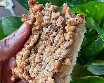 May include: A close-up of a pecan pie bar with a golden brown crust and a generous layer of pecans. The bar is held in a hand and is surrounded by green leaves.
