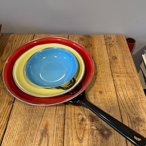 Könnte beinhalten: Ein Satz bunter Emaille-Schüsseln und eine schwarze Bratpfanne auf einer Holzoberfläche. Die Schüsseln sind rot, gelb und blau, mit silberner Umrandung. Die Bratpfanne hat einen langen, schwarzen Griff.