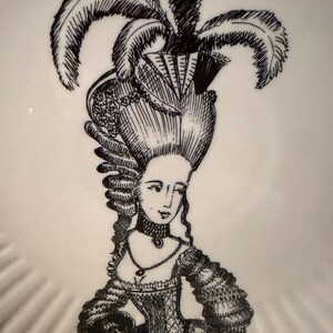 Puede incluir: Un plato blanco con una ilustraci&oacute;n en negro de una mujer con un vestido hist&oacute;rico y un peinado elaborado, adornado con plumas. El plato tiene un borde festoneado y un estilo de dibujo detallado.