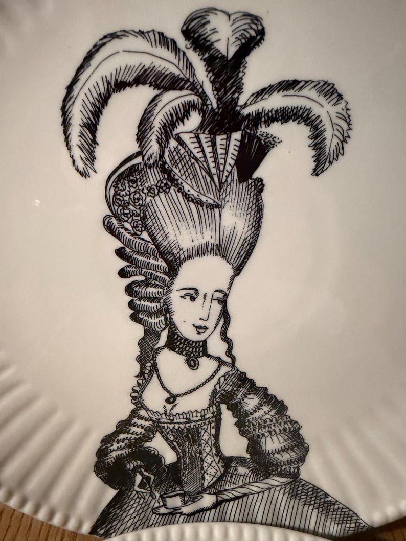 Puede incluir: Un plato blanco con una ilustraci&oacute;n en negro de una mujer con un vestido hist&oacute;rico y un peinado elaborado, adornado con plumas. El plato tiene un borde festoneado y un estilo de dibujo detallado.