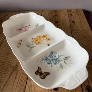 Può includere: Un piatto da portata bianco a tre scomparti con bordo smerlato. Ogni sezione presenta un motivo floreale con farfalle e insetti. I fiori sono nelle tonalità del rosa, giallo e blu. Il piatto è su una superficie di legno.