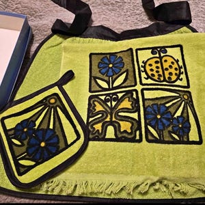 May include: A vintage kitchen set with a light green towel and matching pot holder. The towel has a black border and a fringe. The towel and pot holder are decorated with square designs of flowers, a ladybird, and a butterfly.