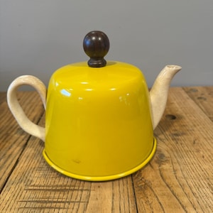 Puede incluir: Una tetera de color amarillo brillante con un cuerpo redondeado, un asa de color claro y un pico. La tapa está rematada con un pomo de madera marrón oscuro. La tetera está sobre una superficie de madera.