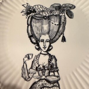 Puede incluir: Plato de cer&aacute;mica blanco con una ilustraci&oacute;n en blanco y negro de una mujer con un vestido hist&oacute;rico y un peinado elaborado, sosteniendo una taza y un platillo. El plato tiene un borde acanalado.