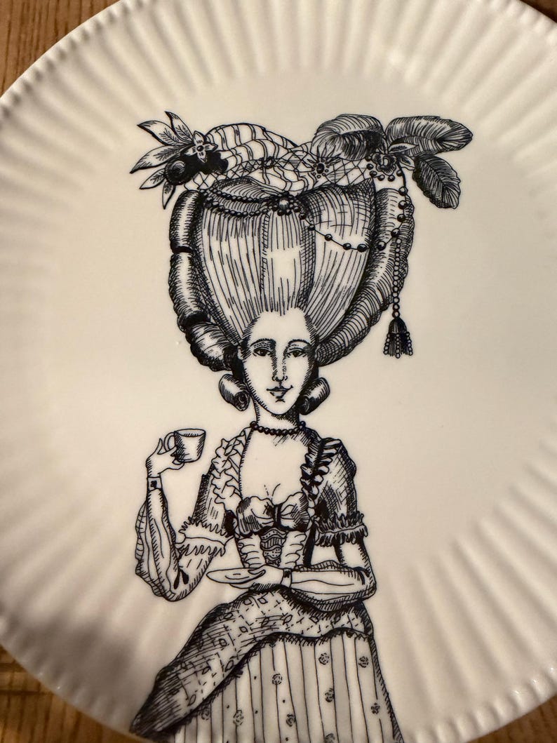 Puede incluir: Plato de cer&aacute;mica blanco con una ilustraci&oacute;n en blanco y negro de una mujer con un vestido hist&oacute;rico y un peinado elaborado, sosteniendo una taza y un platillo. El plato tiene un borde acanalado.