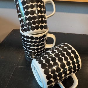 May include: Four white ceramic mugs with a black polka dot pattern. Three mugs are stacked, and one is lying on its side. Each mug has a white handle. The mugs are on a dark surface.