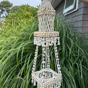 Op de afbeelding: Een hangende schelpenplanter met een gelaagd ontwerp. De planter is gemaakt van witte en bruine schelpen, met een vierkante basis en trapsgewijze strengen schelpen. Het bovenste gedeelte heeft een conische vorm.