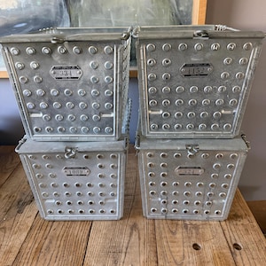 May include: Four vintage metal storage bins with riveted construction and hinged lids. Each bin has a small metal plate with a number. The bins are silver and sit on a wooden surface.