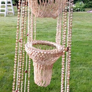 Op de afbeelding: Hangende decoratie van schelpen. Het stuk is gemaakt van kleine, lichtgekleurde schelpen en kralen, gerangschikt in een gelaagd ontwerp. Het bovenste gedeelte heeft een conische vorm, met hangende strengen.