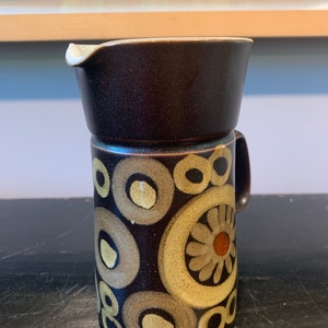 May include: A brown ceramic creamer with a handle and a spout. The creamer is decorated with a pattern of beige and brown circles and flower shapes. The creamer is approximately 5 inches tall.