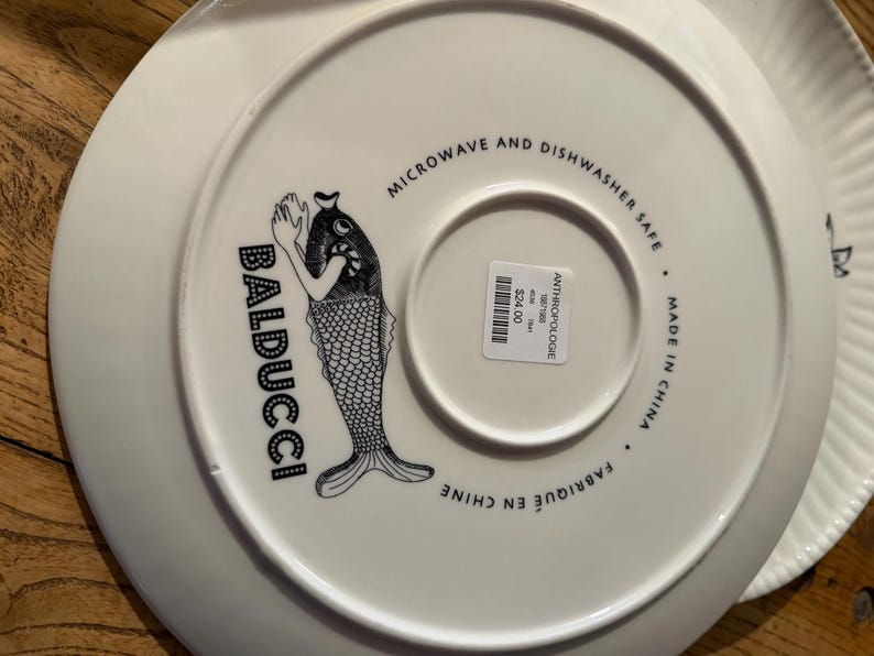 Puede incluir: Plato de cer&aacute;mica blanco con una ilustraci&oacute;n de pez en blanco y negro y la palabra "BADUCCI". El plato est&aacute; etiquetado como "MICROONDAS Y LAVAVAJILLAS" y "MADE IN CHINA".