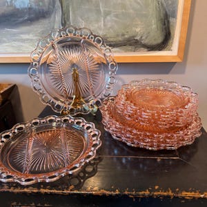 Puede incluir: Un juego de seis platos de servir divididos de vidrio rosa con bordes festoneados. Los platos están apilados sobre una superficie de madera oscura.