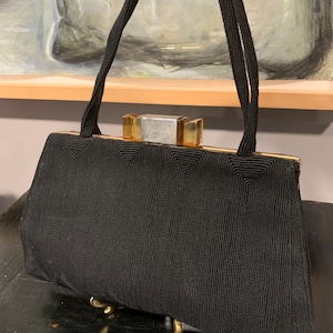 Könnte beinhalten: Eine schwarze Perlenhandtasche mit einem goldenen Verschluss und einem geometrischen Muster. Die Tasche hat zwei lange Henkel.