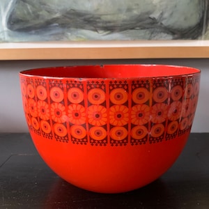 Puede incluir: Un tazón mezclador grande de color naranja brillante con un patrón geométrico en tonos de marrón, naranja y rojo. El patrón presenta círculos, diamantes y flores estilizadas.