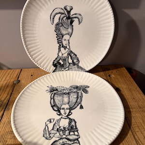 Puede incluir: Dos platos de cer&aacute;mica blanca con borde acanalado. Cada plato presenta una ilustraci&oacute;n en blanco y negro de una mujer con un vestido hist&oacute;rico y un peinado elaborado. Los platos son probablemente para uso decorativo o de comedor.