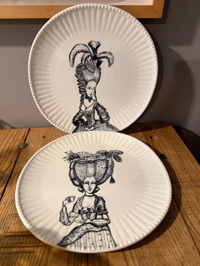 Puede incluir: Dos platos de cer&aacute;mica blanca con borde acanalado. Cada plato presenta una ilustraci&oacute;n en blanco y negro de una mujer con un vestido hist&oacute;rico y un peinado elaborado. Los platos son probablemente para uso decorativo o de comedor.