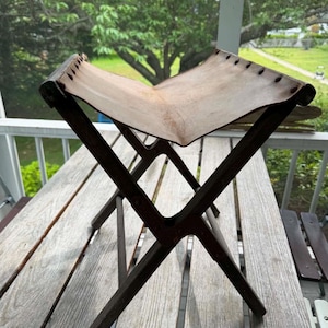 May include: A folding stool featuring a dark brown wooden X-frame base and a light tan leather seat. The leather seat is secured to the wooden frame with metal rivets along the top edges, creating a curved, suspended surface for sitting.
