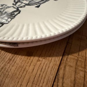 Puede incluir: Dos platos de cer&aacute;mica blanca apilados. Cada plato presenta una ilustraci&oacute;n en blanco y negro de una mujer con vestimenta hist&oacute;rica. Los platos tienen un dise&ntilde;o de borde acanalado. Sobre una superficie de madera.