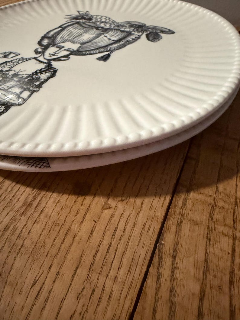 Puede incluir: Dos platos de cer&aacute;mica blanca apilados. Cada plato presenta una ilustraci&oacute;n en blanco y negro de una mujer con vestimenta hist&oacute;rica. Los platos tienen un dise&ntilde;o de borde acanalado. Sobre una superficie de madera.