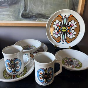 May include: A white ceramic dish set with a retro floral design in brown, orange, blue, and green. The set includes mugs, saucers, bowls, and plates. The design features a stylized floral pattern.