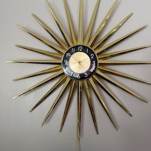 May include: A gold-toned starburst clock with a black and gold face. The clock has long, pointed rays extending outward from the central clock face. The clock face displays the time with gold hands and numerals.