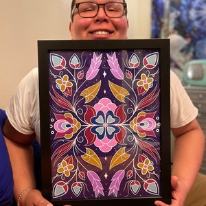 Puede incluir: Una obra de arte enmarcada con un diseño simétrico que presenta flores y hojas estilizadas en tonos de morado, rosa, rojo, dorado y azul sobre un fondo morado oscuro.