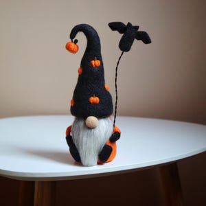 May include: A Halloween gnome doll with a black hat decorated with orange pumpkins. The gnome has a long white beard, an orange body, and holds a black bat. The doll is on a white table.