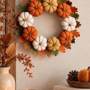 May include: A circular wreath featuring felt pumpkins in orange, white, and yellow, interspersed with felt oak leaves in green and brown. The text A COZY AUTUMN ACCENT FOR YOUR HOME is printed above the wreath with a small maple leaf icon.