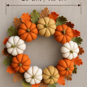 May include: A circular wreath featuring felt pumpkins in orange, white, and tan, interspersed with felt oak leaves in green, orange, and brown. The wreath measures 20 cm in diameter as indicated by the text 20 CM / 7.9 IN above the item.